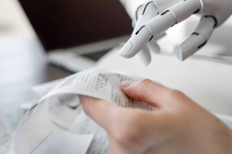 Human hand holding receipt with robot hand pointing at it and receipts and laptop in background